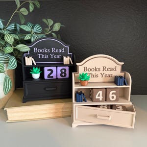 May include: Two miniature bookshelf-style trackers labeled Books Read This Year. One is black with purple number blocks and a green succulent, the other is beige with brown number blocks, blue book accents, and a green succulent in a brown pot.