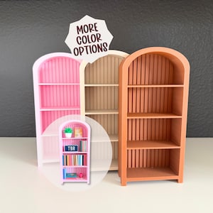 May include: Three miniature bookshelves in pink, beige, and orange. The orange bookshelf is in the foreground, with a curved top and vertical grooves. A smaller pink bookshelf with books is in a circular inset. The text "MORE COLOR OPTIONS" is at the top.