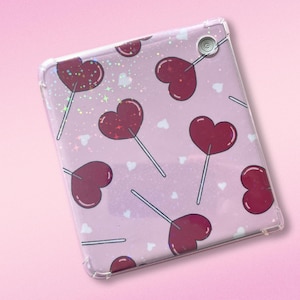 May include: A pink and clear phone case with a repeating pattern of red heart-shaped lollipops on a sparkly background.