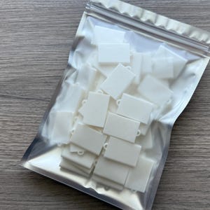 May include: A clear plastic resealable pouch filled with a collection of small, white, rectangular charms. Each charm features a smooth surface and a single circular loop at the top, designed for attachment to jewelry or other craft projects.
