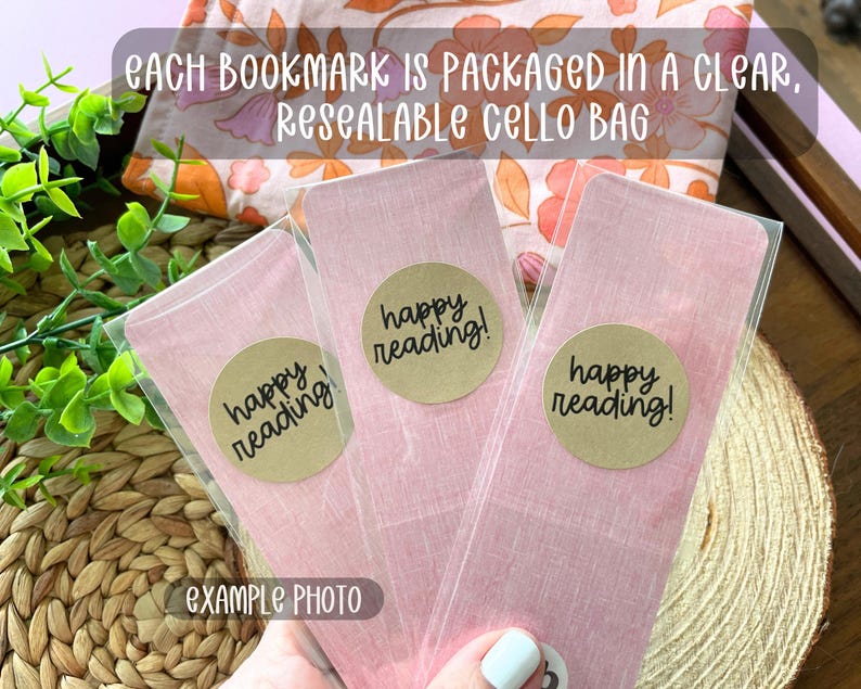 May include: Three rectangular pink bookmarks with a subtle linen-like texture, each enclosed in a clear, resealable cello bag. A round tan sticker is affixed to the front of each bag, featuring the black printed text "happy reading!".