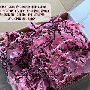 May include: An open cardboard shipping box filled with crinkled paper filler in shades of light pink and dark burgundy. A white text box overlay reads: EVERY ORDER IS PACKED WITH EXTRA CARE BECAUSE I BELIEVE SHOPPING SMALL SHOULD FEEL SPECIAL THE MOMENT YOU OPEN YOUR BOX!