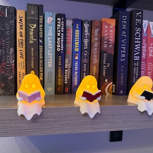 May include: Three small, glowing, yellow ghost-shaped figurines are perched on the edge of a shelf in front of a row of books. Each ghost has two eye cutouts and is holding a small, open book in its hands, colored light purple, red, and black.