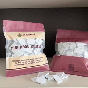 Op de afbeelding: Twee roze hersluitbare zakjes gevuld met kleine witte rechthoekige boekblanco's. Het linkerzakje bevat de tekst "graciebird BOUTIQUE" en "MINI BOOK BLANKS" met beschrijvende tekst. Het rechterzakje toont een QR-code en een website.