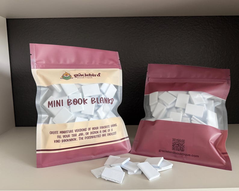 Op de afbeelding: Twee roze hersluitbare zakjes gevuld met kleine witte rechthoekige boekblanco's. Het linkerzakje bevat de tekst "graciebird BOUTIQUE" en "MINI BOOK BLANKS" met beschrijvende tekst. Het rechterzakje toont een QR-code en een website.
