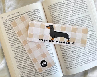 Puede incluir: Dos marcapáginas rectangulares de papel con un patrón de cuadros beige y blanco. Uno muestra una ilustración de un perro salchicha negro y fuego con el texto are you reading smut again? El otro tiene un logotipo circular negro con las letras gb.