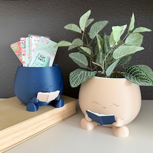May include: Two whimsical, round planters, one navy blue and one beige, each with a face and holding a book. The blue planter holds colorful cards. The beige planter contains a green plant with white-veined leaves. Both are on a book.