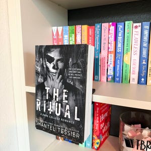 May include: A paperback book titled The Ritual by Shantel Tessier with a black and white cover featuring a person with a skull mask. The book is displayed on a white shelf in front of a row of various colorful books and a glass jar labeled TBR.