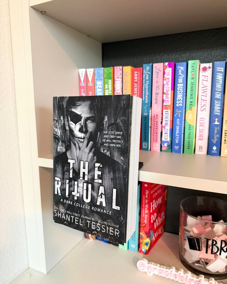 May include: A paperback book titled The Ritual by Shantel Tessier with a black and white cover featuring a person with a skull mask. The book is displayed on a white shelf in front of a row of various colorful books and a glass jar labeled TBR.
