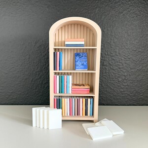 Puede incluir: Una estanter&iacute;a de madera en miniatura con una parte superior arqueada, llena de libros coloridos. La estanter&iacute;a es de color claro y los libros tienen varios colores. Varios bloques rectangulares blancos est&aacute;n frente a la estanter&iacute;a.