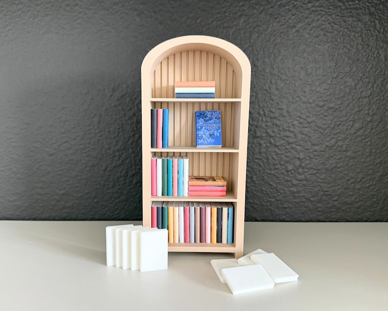 Puede incluir: Una estanter&iacute;a de madera en miniatura con una parte superior arqueada, llena de libros coloridos. La estanter&iacute;a es de color claro y los libros tienen varios colores. Varios bloques rectangulares blancos est&aacute;n frente a la estanter&iacute;a.