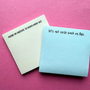 May include: Two square sticky notes, one pale yellow and one light blue. The yellow note features the black printed text 'PLEASE DO HESITATE TO REACH BACK OUT' and the blue note features the black printed text 'let's not circle back on this...'