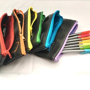 May include: A set of six colourful zippered pouches made from recycled inner tubes. The pouches are lined with different colours, including red, orange, yellow, green, blue, and purple. The pouches are arranged in a rainbow pattern and are filled with colourful pens.