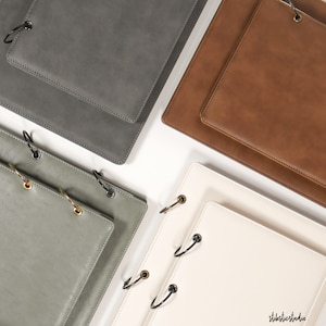 Puede incluir: Cuatro cuadernos de diferentes tamaños encuadernados en cuero con anillos metálicos. Los cuadernos son en tonos de gris, verde, marrón y blanco.