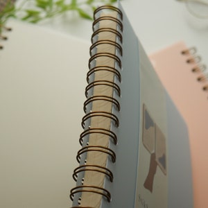 May include: Close-up of three spiral-bound notebooks. The notebook in focus has a light blue cover with a brown spiral binding. The pages are white. A second notebook is pink, and a third is white. The background is white.