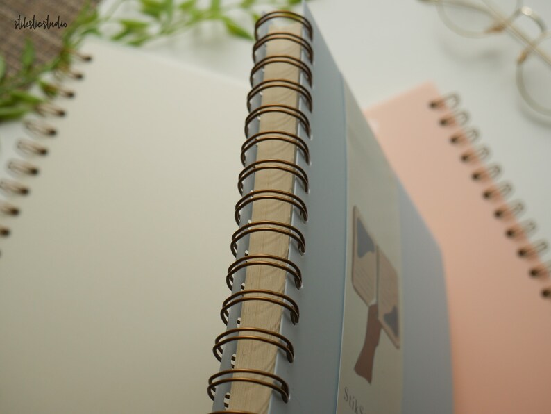 May include: Close-up of three spiral-bound notebooks. The notebook in focus has a light blue cover with a brown spiral binding. The pages are white. A second notebook is pink, and a third is white. The background is white.