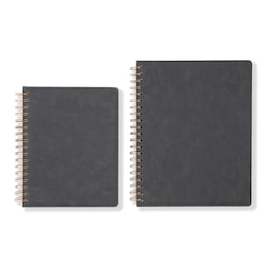 May include: Two charcoal grey spiral-bound notebooks. The notebooks have a textured cover and a gold-coloured spiral binding. One notebook is smaller and square, the other is larger and rectangular.
