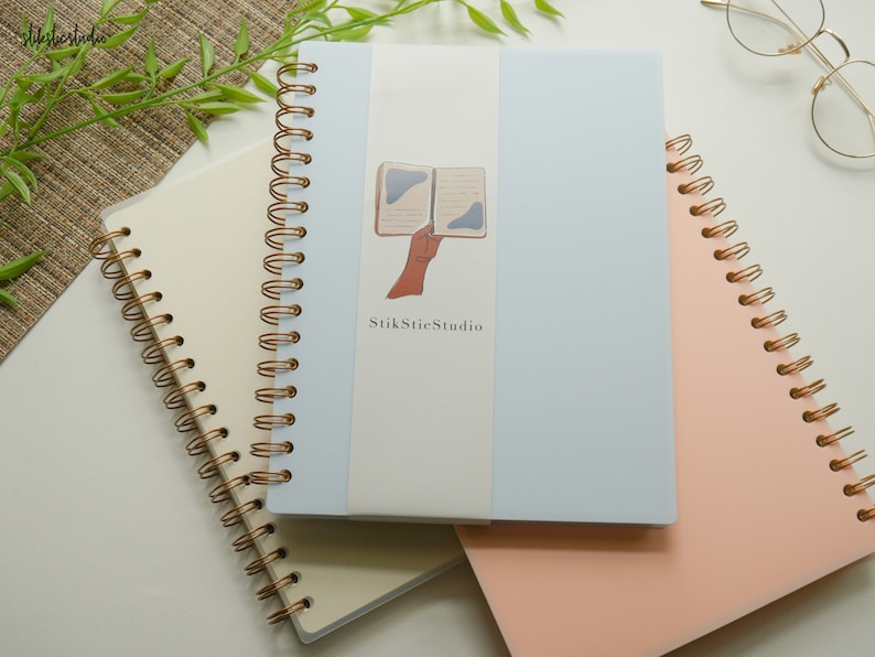 May include: Three spiral-bound notebooks in cream, light blue, and peach. The light blue notebook has a sticker with a hand holding a book and the text "StikStieStudio". Gold-colored spiral binding is visible on each notebook.