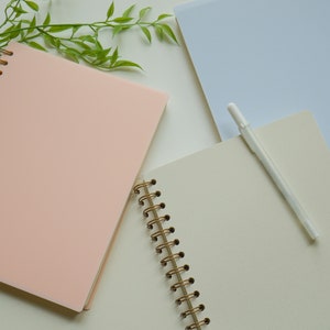 May include: An overhead shot of three spiral-bound notebooks in pastel colors: light pink, light blue, and cream. A white pen rests on the open cream-colored notebook. The notebooks have gold-colored spiral bindings. Green leafy sprigs are in the background.