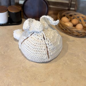 May include: A white crocheted bunny hat with a pom-pom on top. The hat is folded in half and the ears are sticking up.