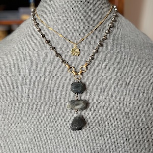 May include: A layered necklace set on a gray mannequin. The top necklace is gold with a small lotus flower pendant. The bottom necklace has dark gray beads and a pendant with three stacked, gray stone pieces.