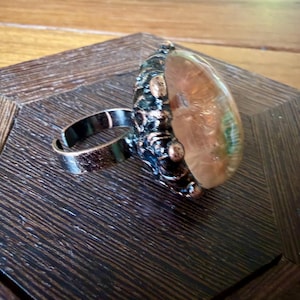May include: An adjustable ring featuring a dark, textured metallic band with a copper-toned finish. The setting holds a large, translucent, rounded cabochon stone that displays swirling amber, peach, and subtle green hues within its smooth surface.
