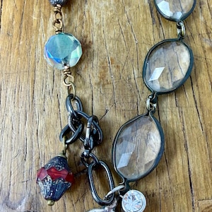 May include: A handcrafted bracelet featuring a mix of beads and charms. The bracelet includes faceted clear and blue beads, red and black decorative beads, and a silver-toned medallion. A small, clear crystal charm hangs near the medallion.