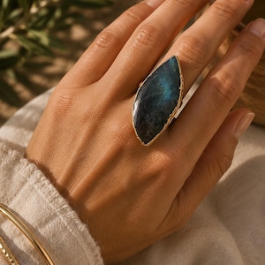 May include: A marquise-shaped labradorite ring featuring a dark blue and grey iridescent stone set in a gold-toned metal band. The ring is worn alongside two thin, gold-toned metal bangles on the wrist, showcasing a smooth, polished finish.