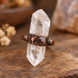 May include: A ring featuring a double-terminated clear quartz crystal point set horizontally within a textured, dark copper-toned metal band. The crystal is transparent and faceted, while the band has a rugged, hammered finish.