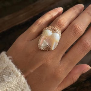 May include: A ring featuring a large, irregular, iridescent white pearl centerpiece set within a textured, rose gold-toned metal band encrusted with numerous small, sparkling, light brown pavé stones that frame the pearl's organic shape.