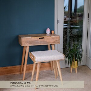 May include: A wooden vanity table with a drawer and a lift-top mirror. The table has a light wood finish and is on tapered legs. A white cushioned stool is in front of the table. The text "PERSONALISE ME" and "AVAILABLE IN 2 SIZES & 14 HANDPAINTED OPTIONS" is below the table.