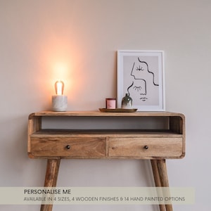 May include: A wooden console table with two drawers and a shelf. The table has a light brown finish and features rounded edges. The table is decorated with a white marble lamp base, a framed black and white line art print, and a small green vase with a pink candle.