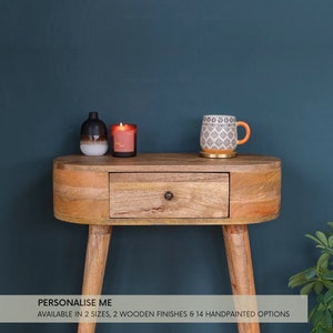 May include: A wooden console table with a single drawer and rounded edges. The table has a natural wood finish and is supported by four turned legs. The table is decorated with a candle, a small vase, and a patterned mug.