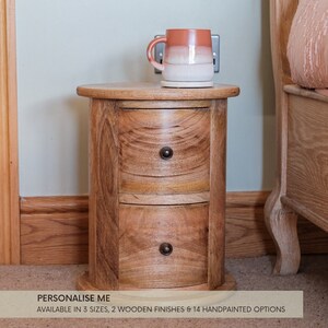May include: A wooden bedside table with two drawers and a rounded top. The table is made of light brown wood and has a natural finish. The table is sitting on a carpeted floor next to a bed. The text "PERSONALISE ME" is visible at the bottom of the image. The text "AVAILABLE IN 3 SIZES, 2 WOODEN FINISHES & 14 HANDPAINTED OPTIONS" is also visible.