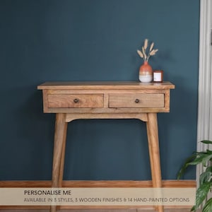 May include: A wooden console table with two drawers and tapered legs. The table is a light brown colour and has a natural wood finish. The table is sitting in front of a teal wall.