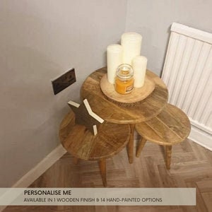 May include: Two wooden nesting tables with a star-shaped ornament on the top table. The tables are in a room with white walls and a white radiator. There are candles and a jar of scented wax on the top table.