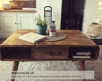 Könnte beinhalten: Ein Holz-Couchtisch mit einer Schublade und zwei offenen Regalen. Der Tisch hat eine natürliche Holzoptik und steht auf einem Teppich. Der Text "PERSONALISE ME" und "AVAILABLE IN 3 WOODEN FINISHES & 14 HAND-PAINTED OPTIONS" ist am unteren Rand des Bildes sichtbar.