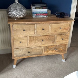 May include: A light brown wooden chest of drawers with various sized drawers. A silver lamp with a white shade and books are on top. The chest has four short, angled legs. A bottle of champagne rests on the floor.