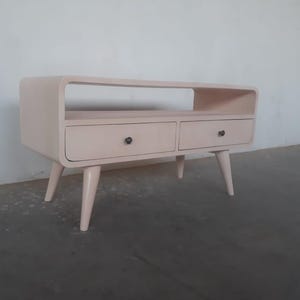 May include: A light pink mid-century modern style TV stand with two drawers and a shelf. The stand has four tapered legs and black metal drawer pulls.