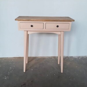 May include: A blush pink console table with two drawers and a wood top. The table has tapered legs and black drawer pulls.