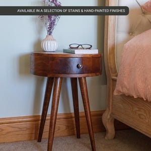 May include: A dark brown, round bedside table with three angled legs. It features a single drawer with a metal knob. A small vase with purple flowers, glasses, and a book are on top. The text reads "AVAILABLE IN A SELECTION OF STAINS & HAND-PAINTED FINISHES." The table is approximately 50 cm in diameter.