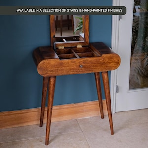 May include: A wooden vanity table with a built-in mirror and storage compartments. The table has a warm brown finish, four tapered legs, and a small drawer. The text "AVAILABLE IN A SELECTION OF STAINS & HAND-PAINTED FINISHES" is displayed at the top.
