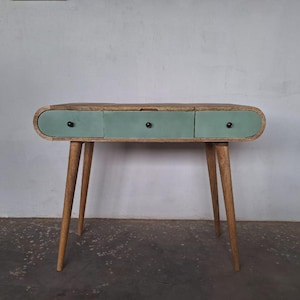 May include: A light green console table with three drawers and rounded edges. The table has a natural wood finish and black knobs on the drawers. The table is on a light grey concrete floor.