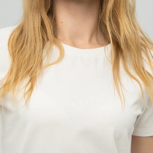 Op de afbeelding: Een wit T-shirt met ronde hals en het woord "MOBA" in reliëf op de borst. Het shirt heeft korte mouwen en een eenvoudig, klassiek ontwerp. Het model heeft blond haar.