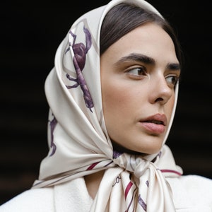 May include: A square silk headscarf in cream featuring a repeating pattern of dark purple horses and thin burgundy and grey stripes. The scarf is tied in a knot under the chin, displaying a smooth, lustrous finish and a classic folded drape.