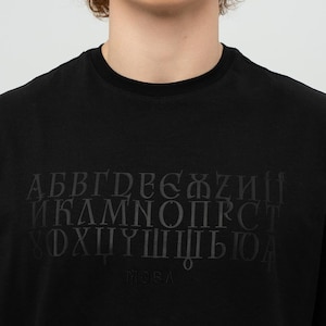 Puede incluir: Camiseta negra de cuello redondo con un diseño de alfabeto cirílico en un tono negro más oscuro. La camiseta presenta la palabra "MOBA" en la parte inferior del diseño. La camiseta está hecha de un material suave.