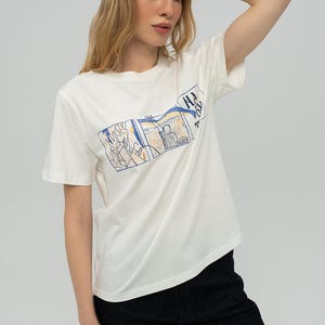 Puede incluir: Camiseta blanca con un estampado gráfico colorido y el texto "NA 20". El gráfico presenta un diseño de paisaje con detalles en azul y amarillo. La modelo lleva vaqueros negros.