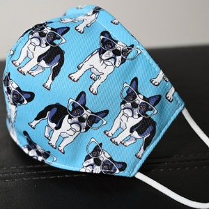 May include: A blue face mask with a white elastic band. The mask has a repeating pattern of black and white French Bulldogs wearing glasses.