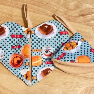 May include: Two heart-shaped pot holders with a blue and white checkered pattern. The pot holders feature images of food and drinks, including pastries, tea, and milk. The text "Black & White Milk" is visible on one of the pot holders.