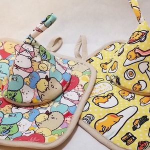May include: Two yellow and white pot holders with a cute cartoon design. One pot holder features a yellow egg character with a white shell and a red yolk. The other pot holder features a variety of cartoon characters with a colorful design. Both pot holders have a beige fabric backing.